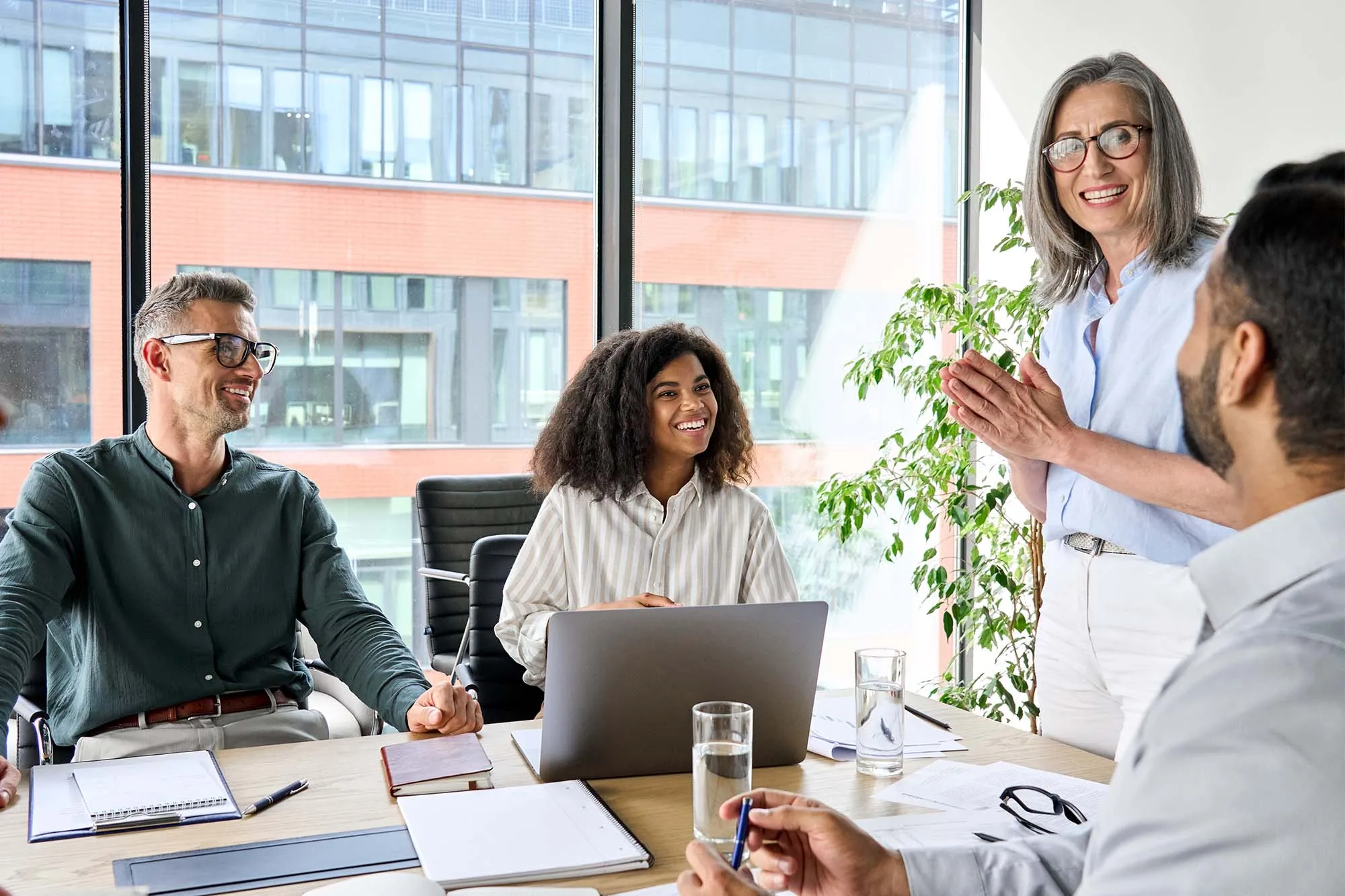 A group of four business people is gathered around a table in a bright office with a large window.