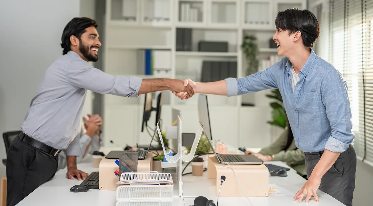Two smiling men, wearing shirts, shake hands across a desk in a modern office
