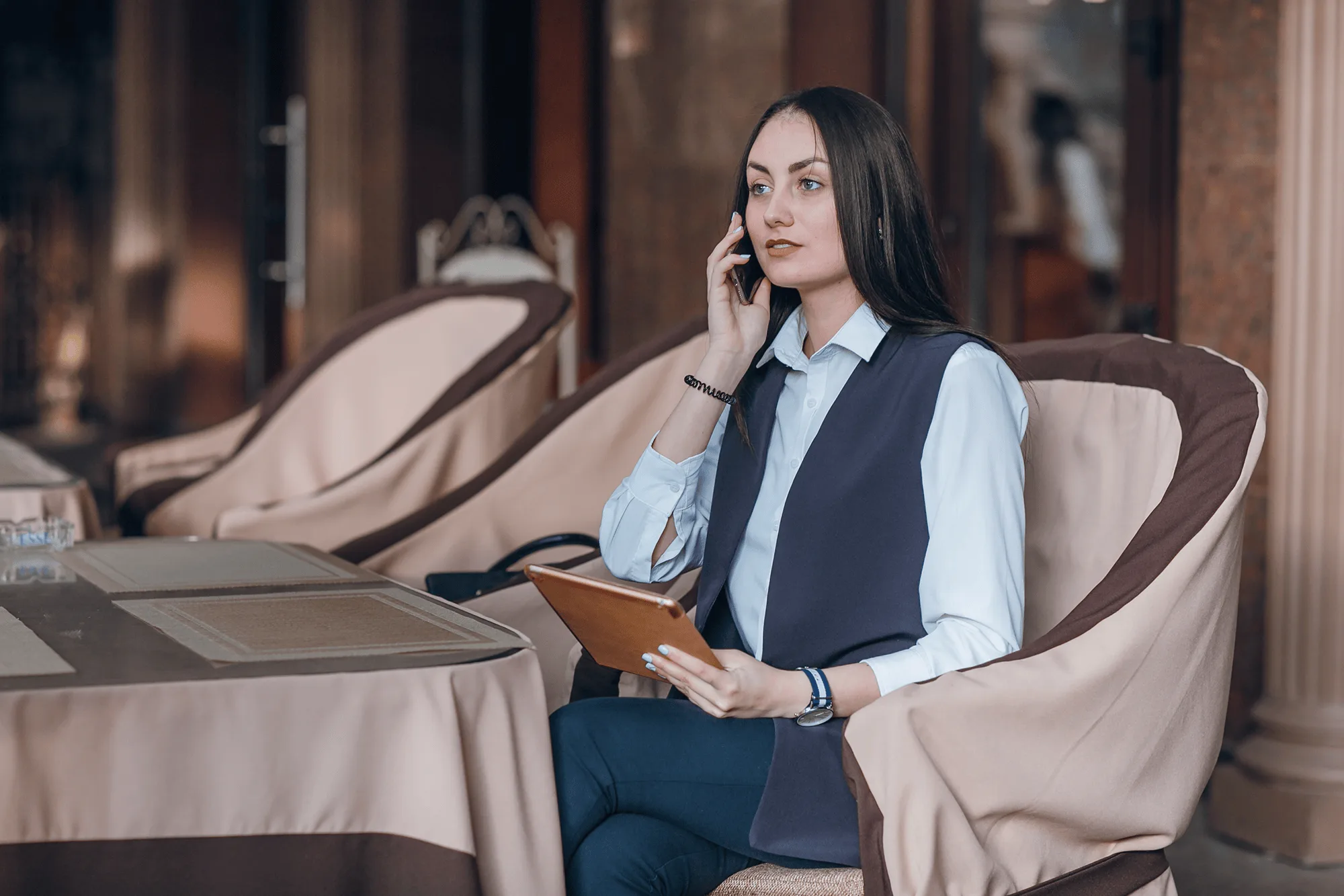 A woman wearing a white collared shirt and a dark vest is talking on her cell phone while holding a tablet in a business setting.