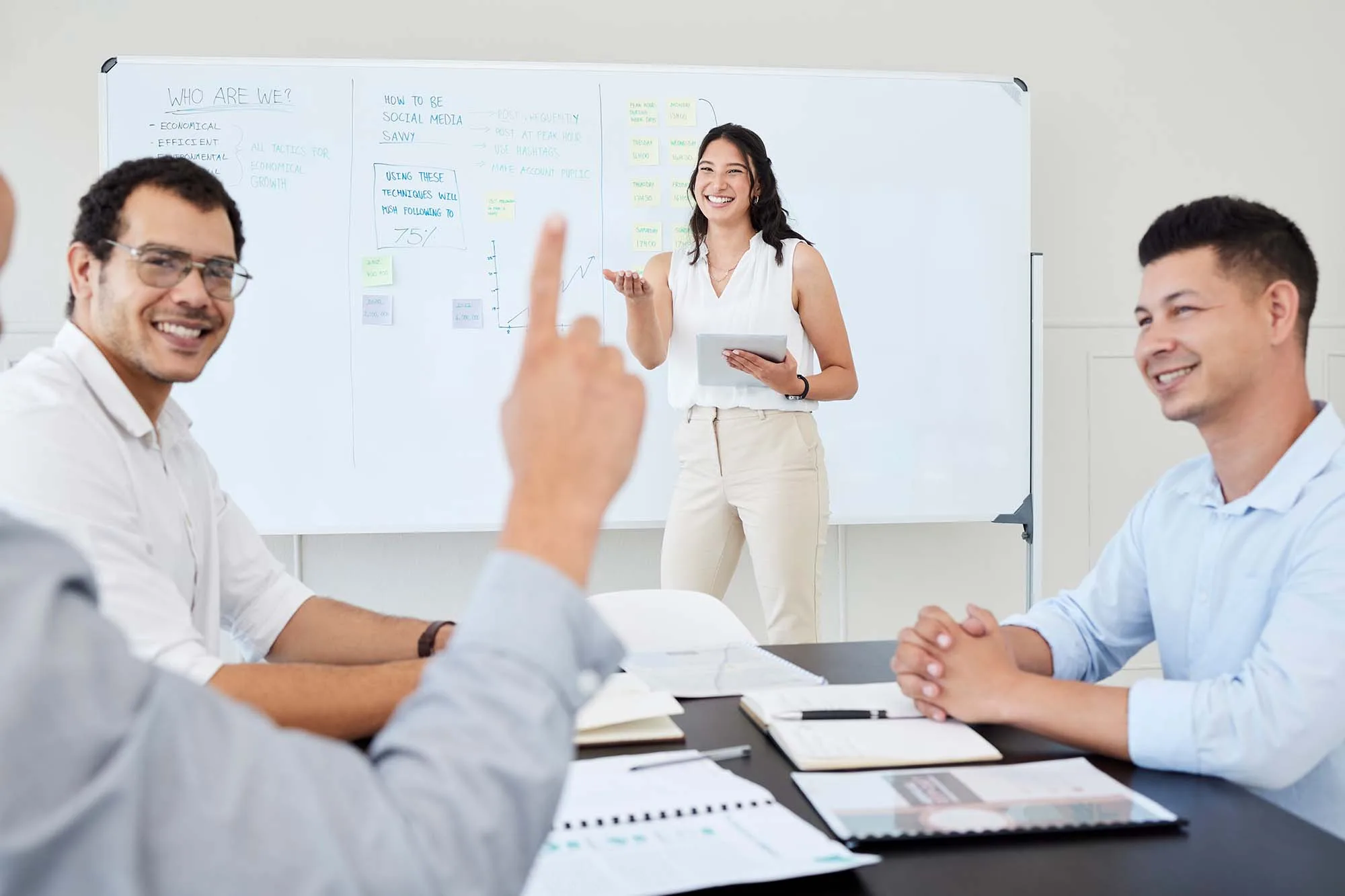 A group of business people are in a meeting, with a woman presenting information from a tablet at a whiteboard