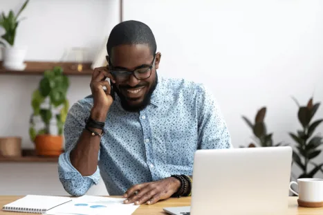 A smiling man wearing glasses is talking on his smartphone while working at a desk with a laptop and charts.