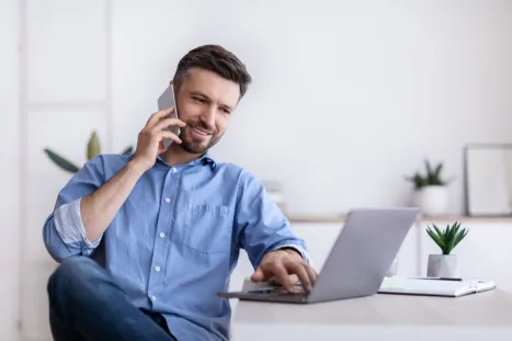A smiling man wearing a blue shirt is sitting at a desk, working on a laptop and talking on his smartphone.