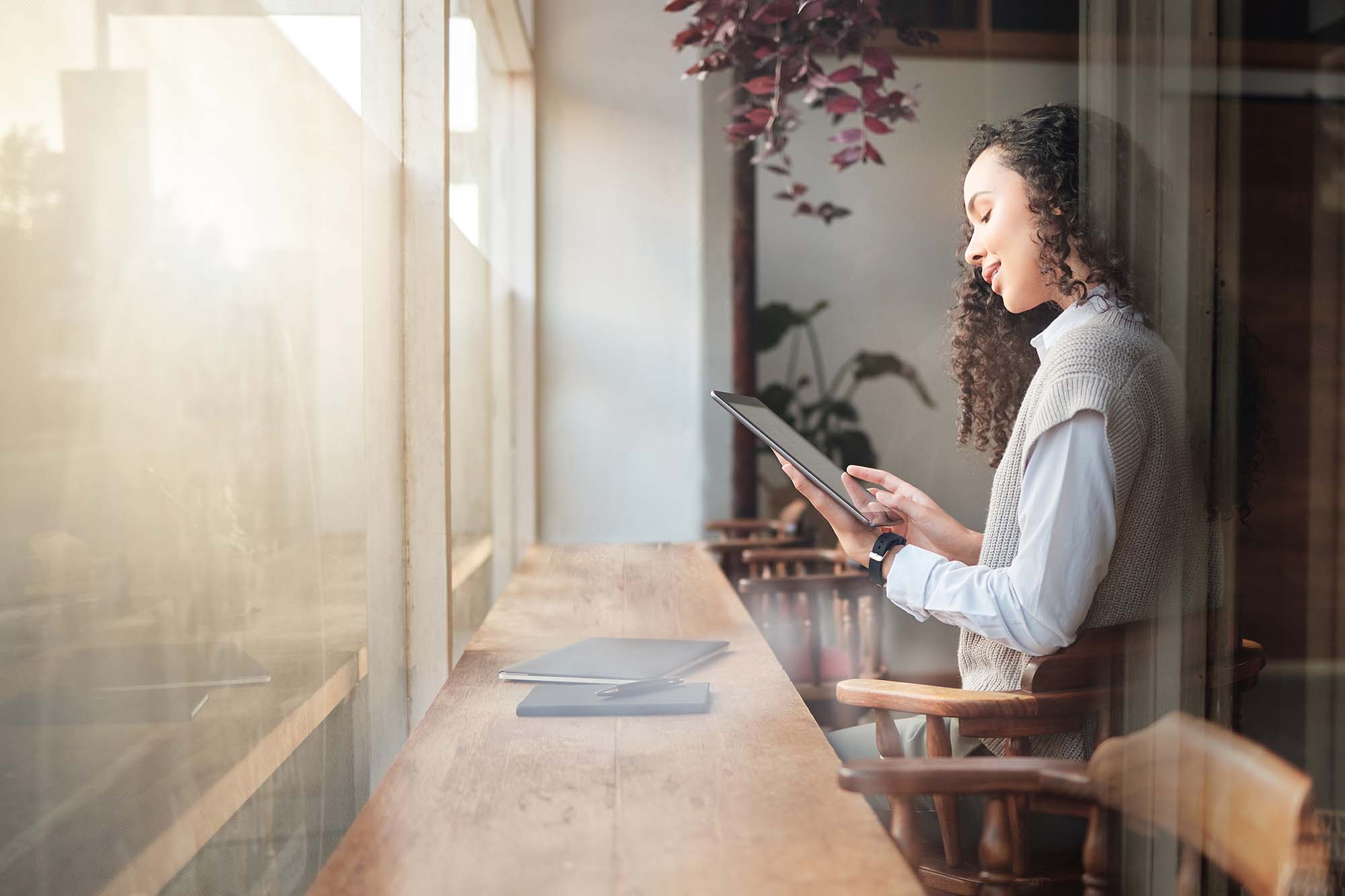 A woman with curly hair is using a tablet while standing next to a window with natural light