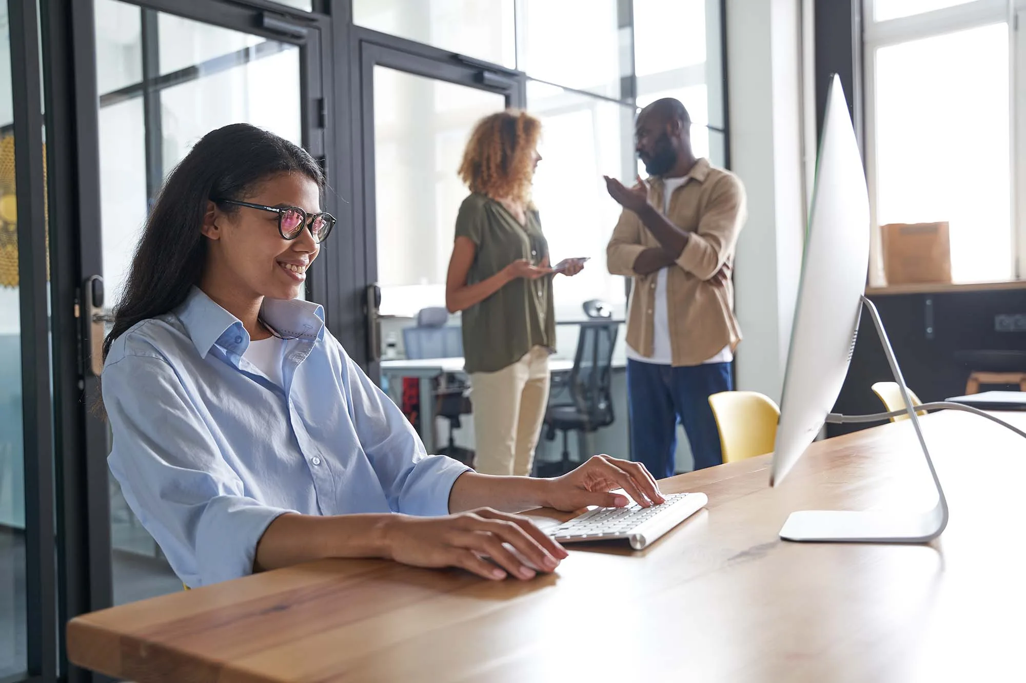 A smiling woman wearing glasses is working at a computer in a bright, modern office setting. Two other coworkers are in the background having a conversation.