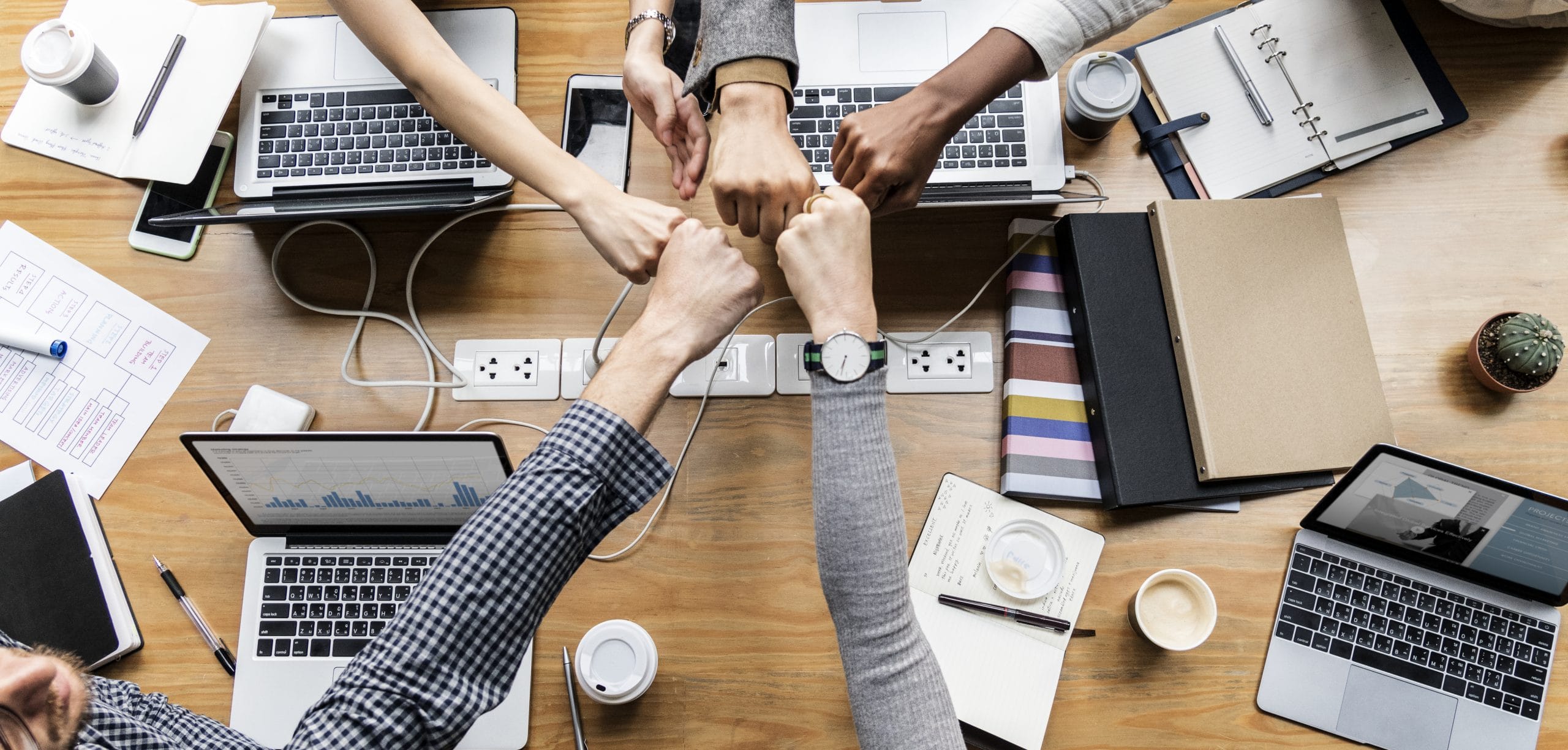Teamwork in the modern office: people's hands come together in a fist bump over a busy workspace with laptops and notebooks