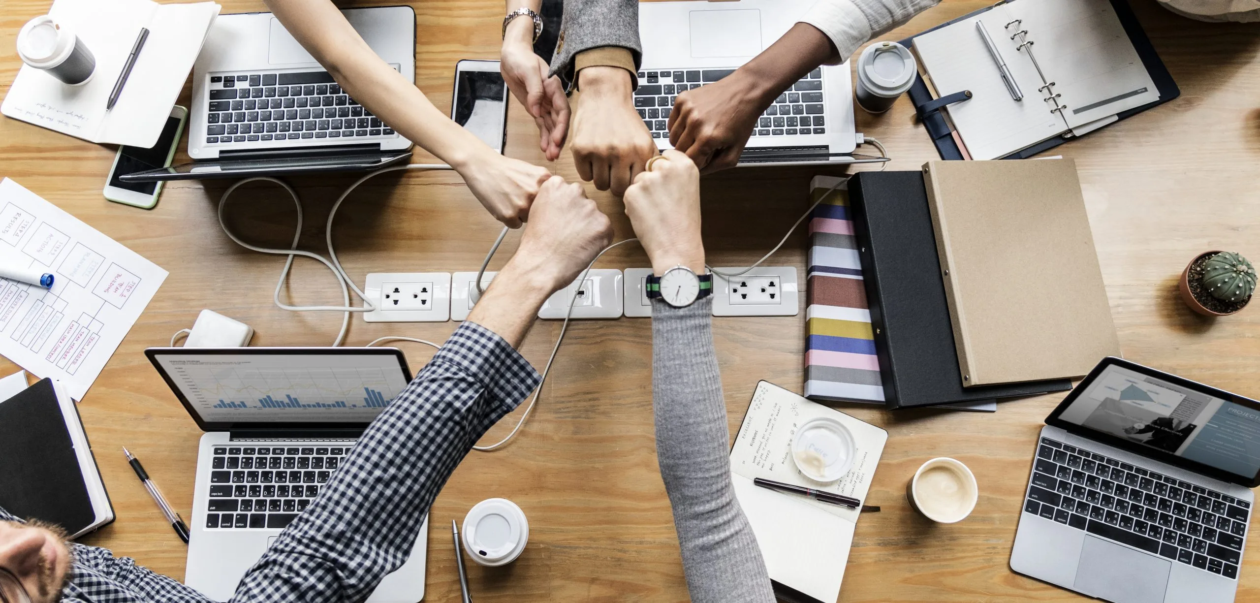 Teamwork in the modern office: people's hands come together in a fist bump over a busy workspace with laptops and notebooks