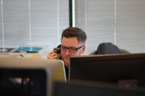 A man wearing glasses is speaking on the phone while working at his desk.