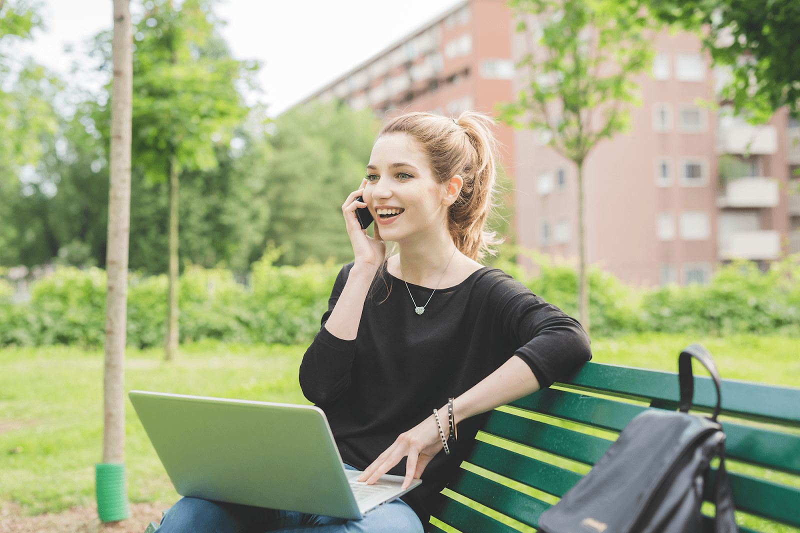 A woman with blonde hair talking on her smartphone, with a laptop on her lap, while sitting on a green park bench.