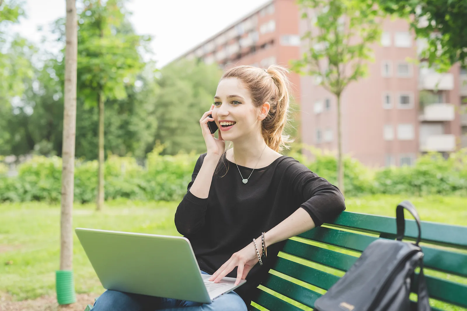 A woman with blonde hair talking on her smartphone, with a laptop on her lap, while sitting on a green park bench.