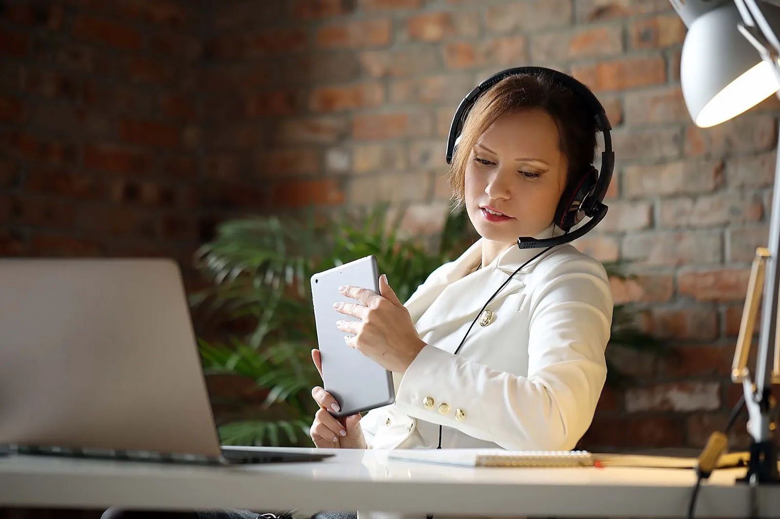 A female customer support representative wearing a white suit jacket and a headset is holding a tablet and working at a desk with a laptop.