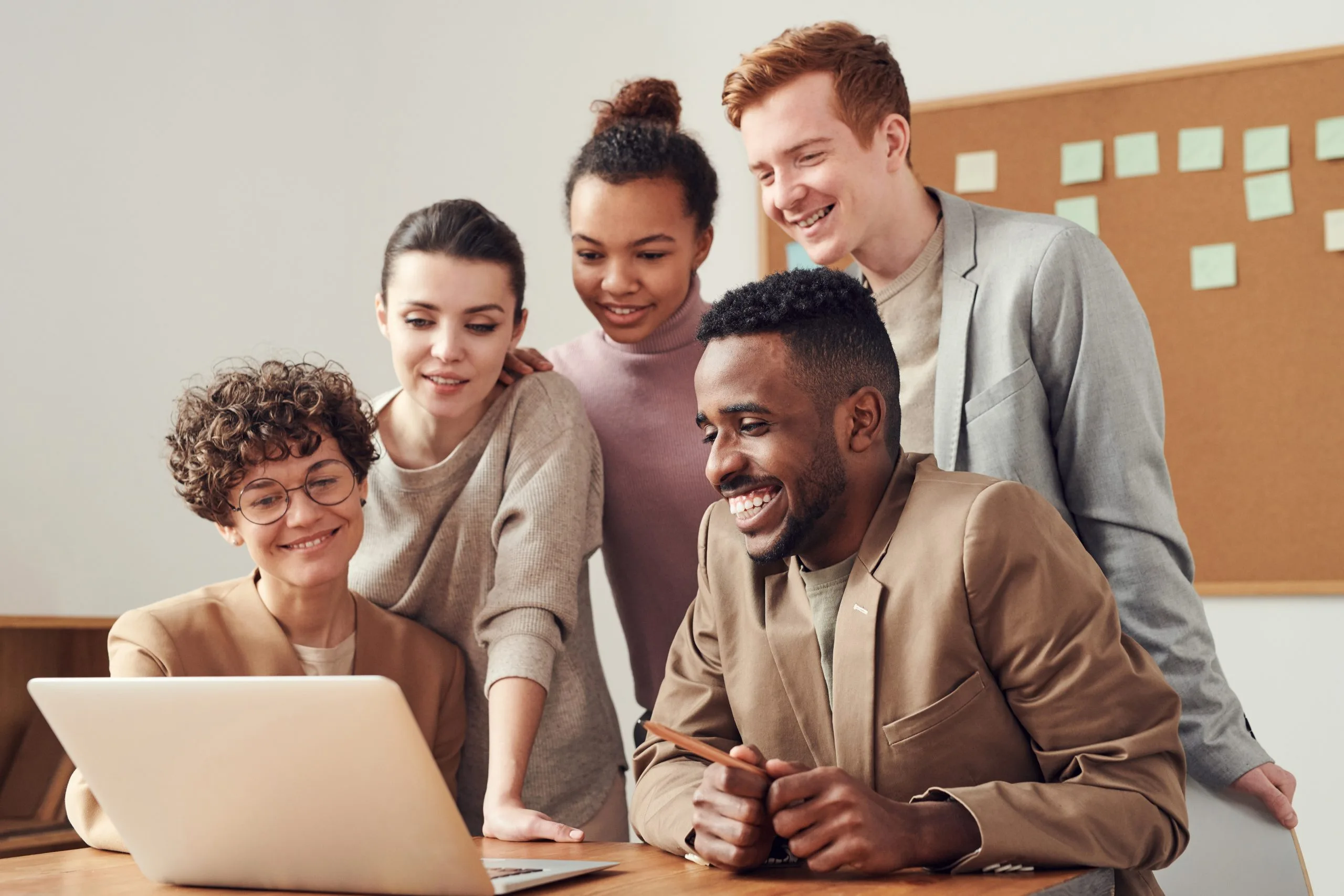 A team of five young professionals gather around a laptop, smiling and engaged in the screen's content. A cork board with sticky notes is in the background
