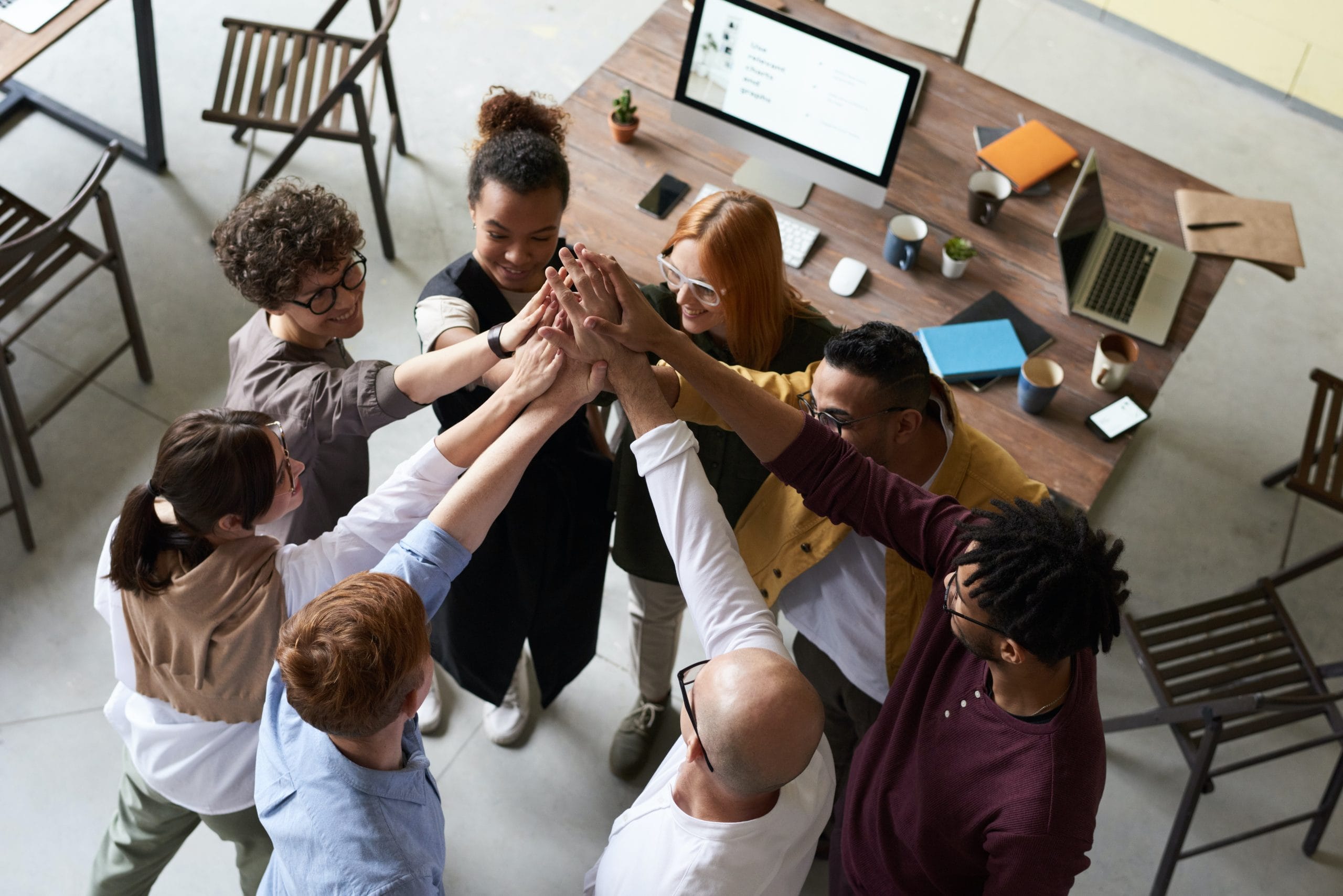 An overhead shot of a team of about 8 people standing in a circle and giving each other a high-five. A large table with a computer, laptops, and other office supplies is in the background