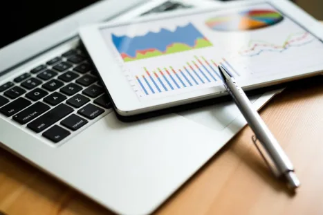 Close-up of a laptop, a tablet displaying business graphs and charts, and a pen on a wooden surface.