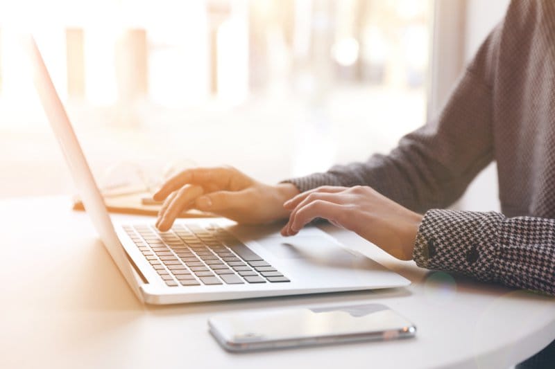 Hands typing on a laptop keyboard with a phone nearby in a brightly lit space.