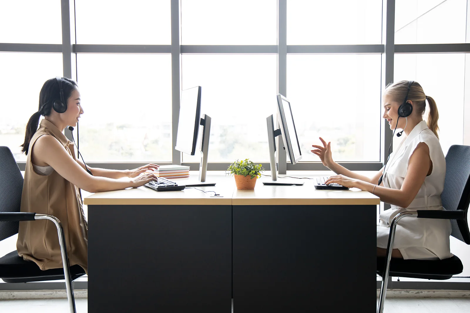 Two women, wearing sleeveless t-shirts and headsets, are sitting opposite each other and working at the computers