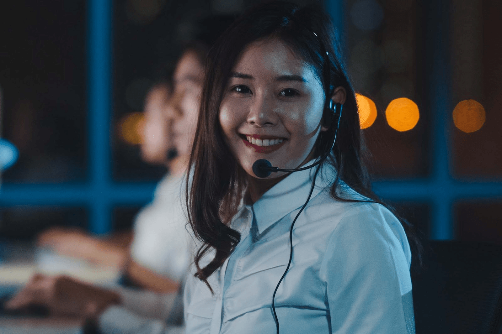 A female customer support representative with long black hair, wearing a white blouse and a headset, is working in a modern office.