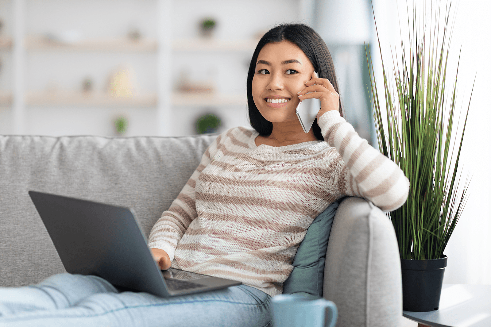 A woman is working remotely from home, sitting on a gray sofa, smiling as she talks on a smartphone and uses a laptop.