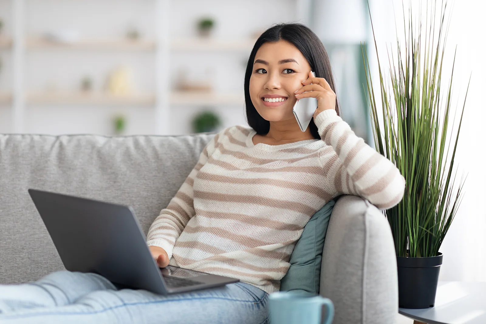 A woman is working remotely from home, sitting on a gray sofa, smiling as she talks on a smartphone and uses a laptop.