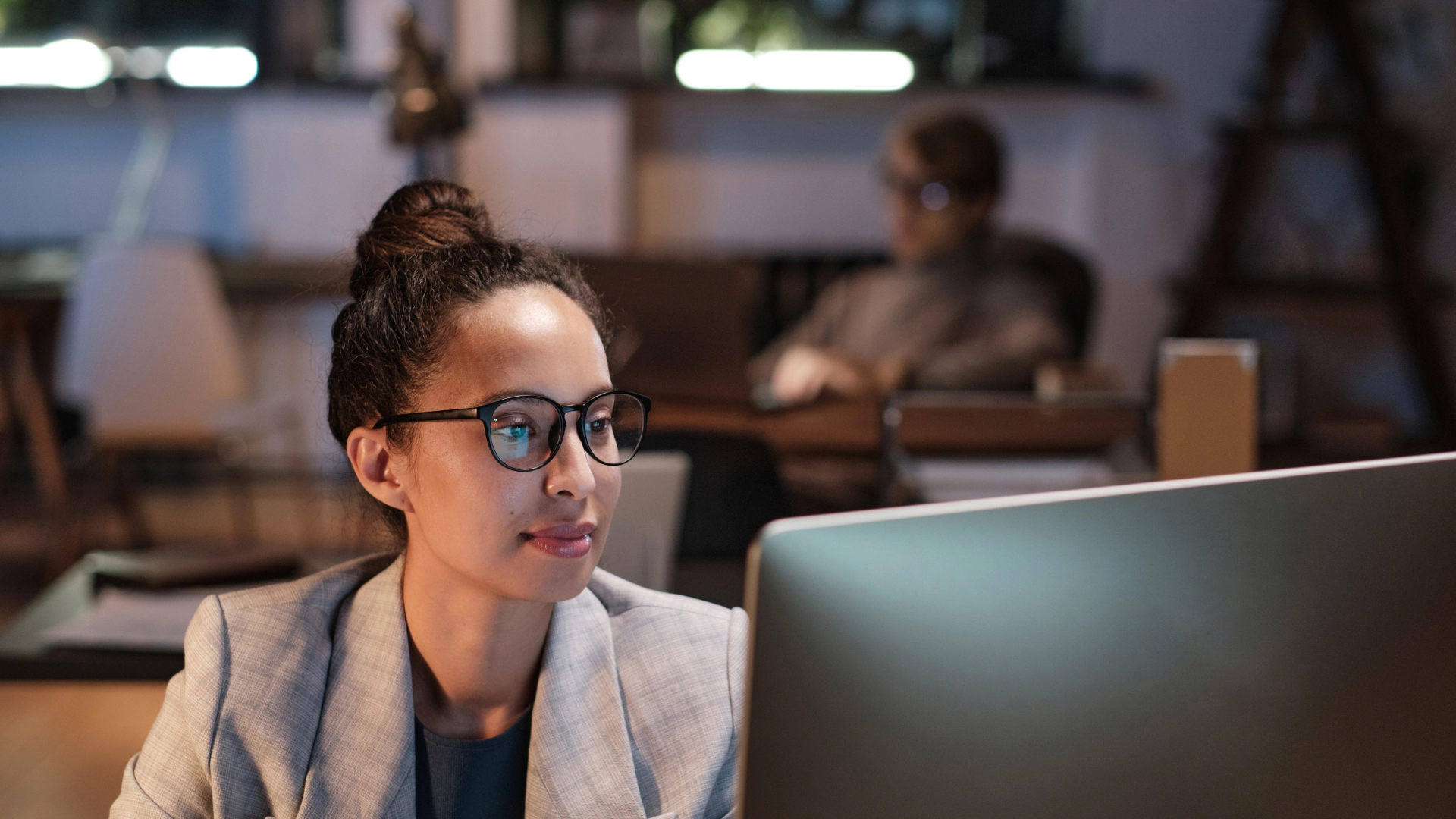 Woman wearing glasses and a blazer, focused on her computer screen in a dimly lit office.