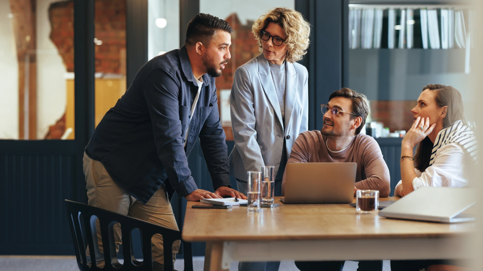Four business colleagues engaged in a meeting, reviewing information and discussing strategies around a table with laptops and notes.