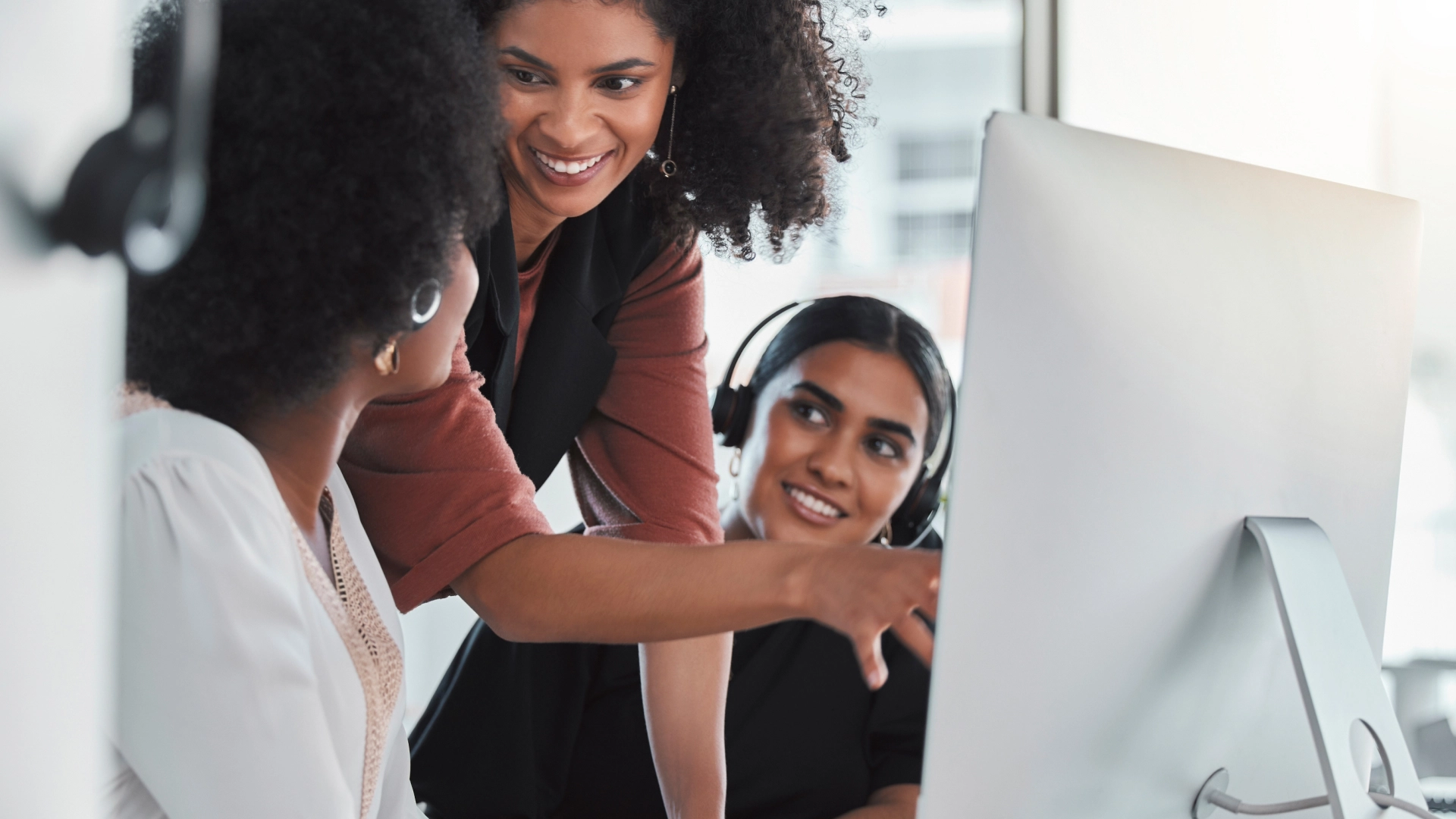 Three women working together on a computer, wearing headsets, represent collaborative teamwork in a customer support role.