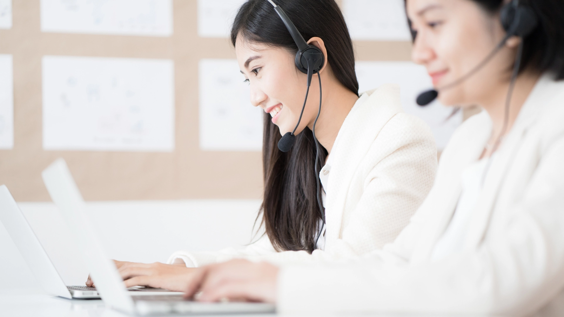 Two smiling women wearing headsets and white blazers, working in a customer service environment.