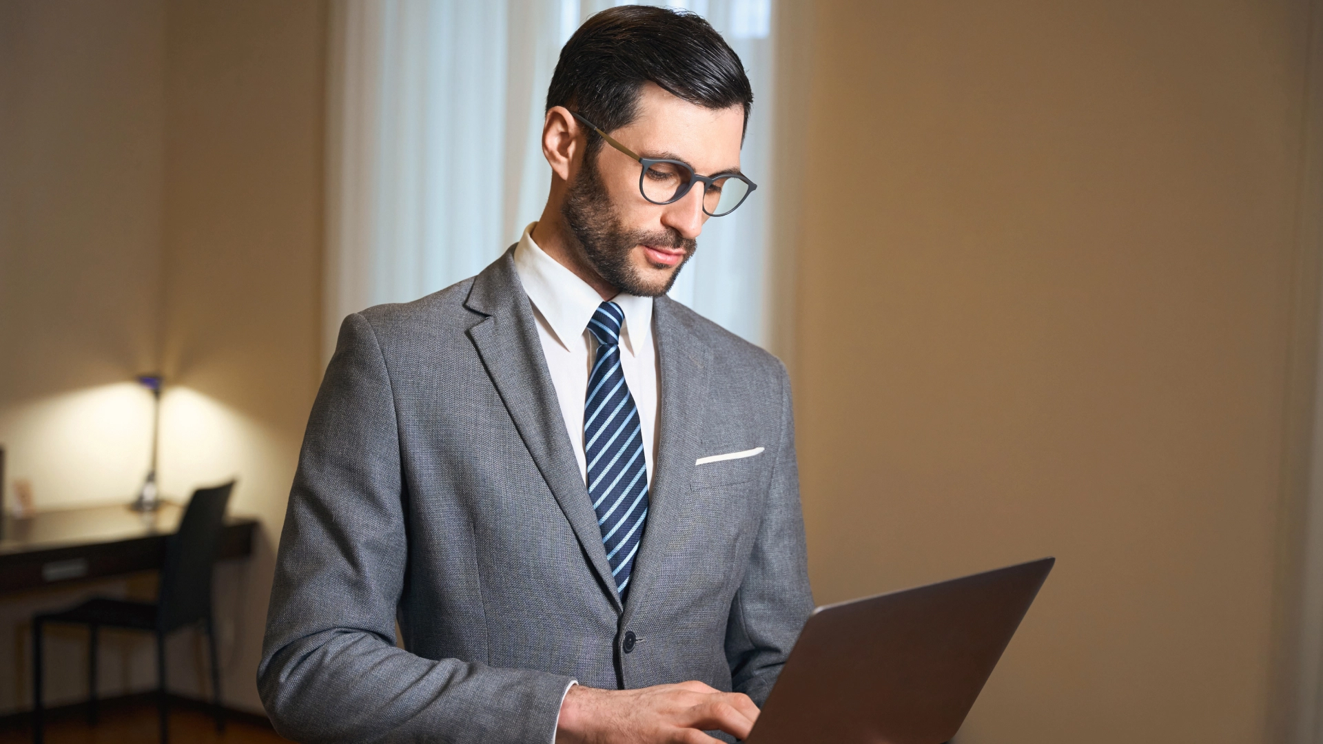 A man with a beard and glasses wearing a grey suit and blue tie, is working on a laptop.