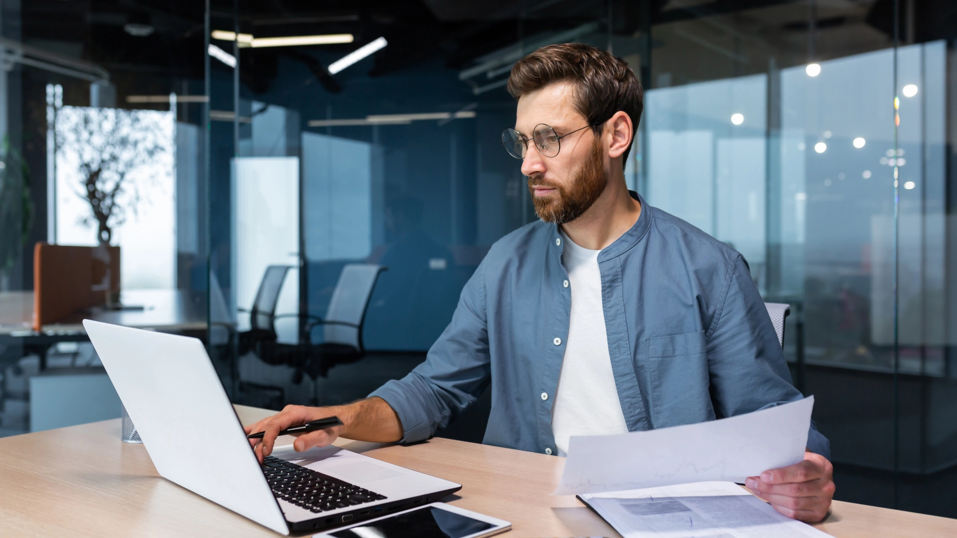 A focused man wearing a blue shirt and glasses works at a laptop in a modern office with glass walls.
