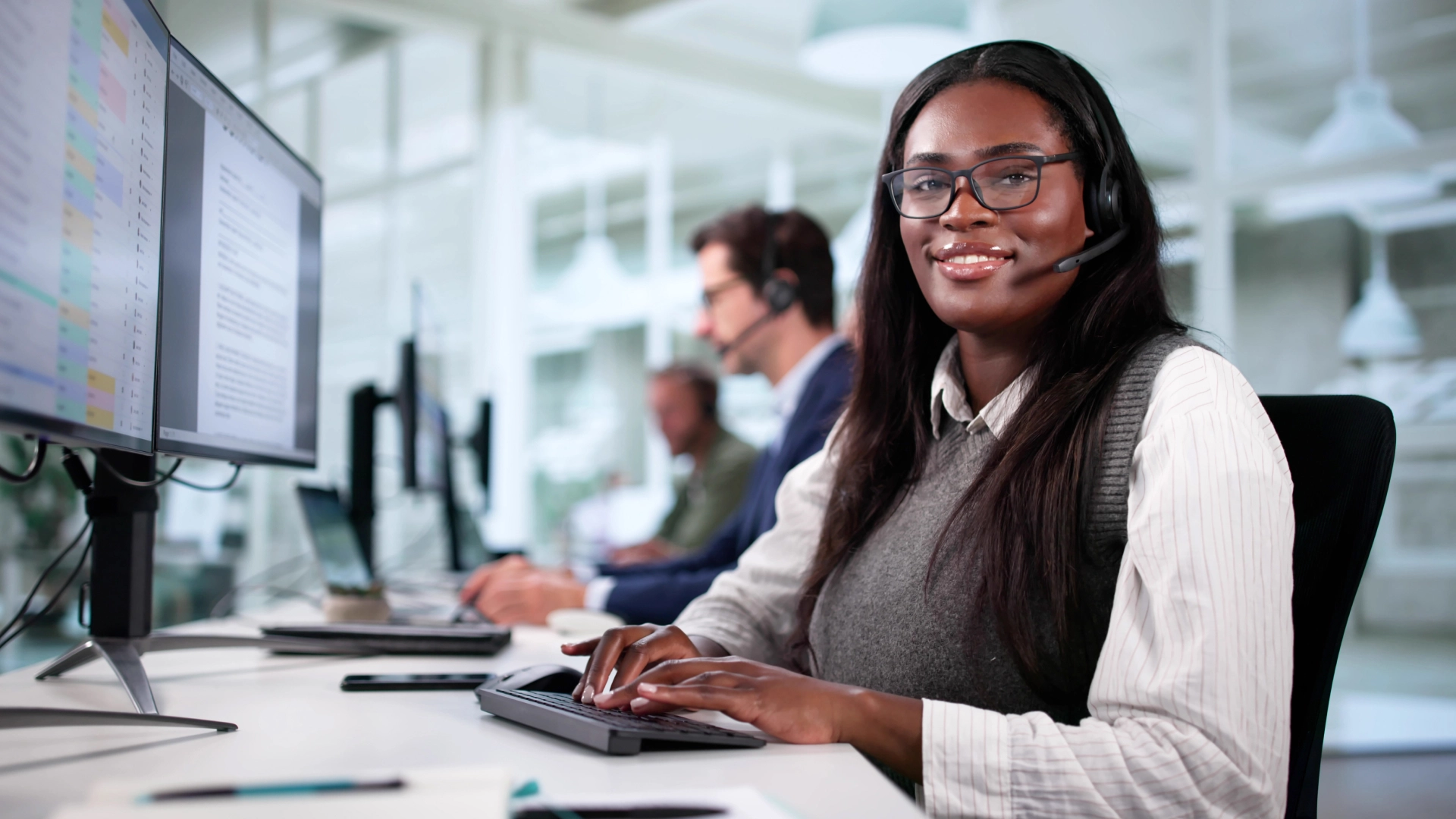 Call center with a smiling woman at the forefront wearing glasses and a headset, with other agents working in the background.
