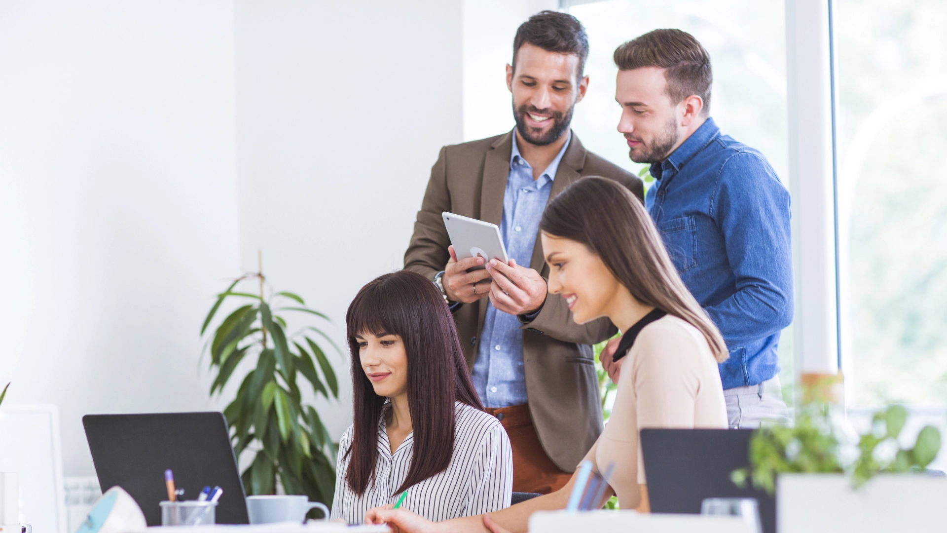 Diverse group of colleagues working together, with a man holding a tablet while two women look at a computer screen.