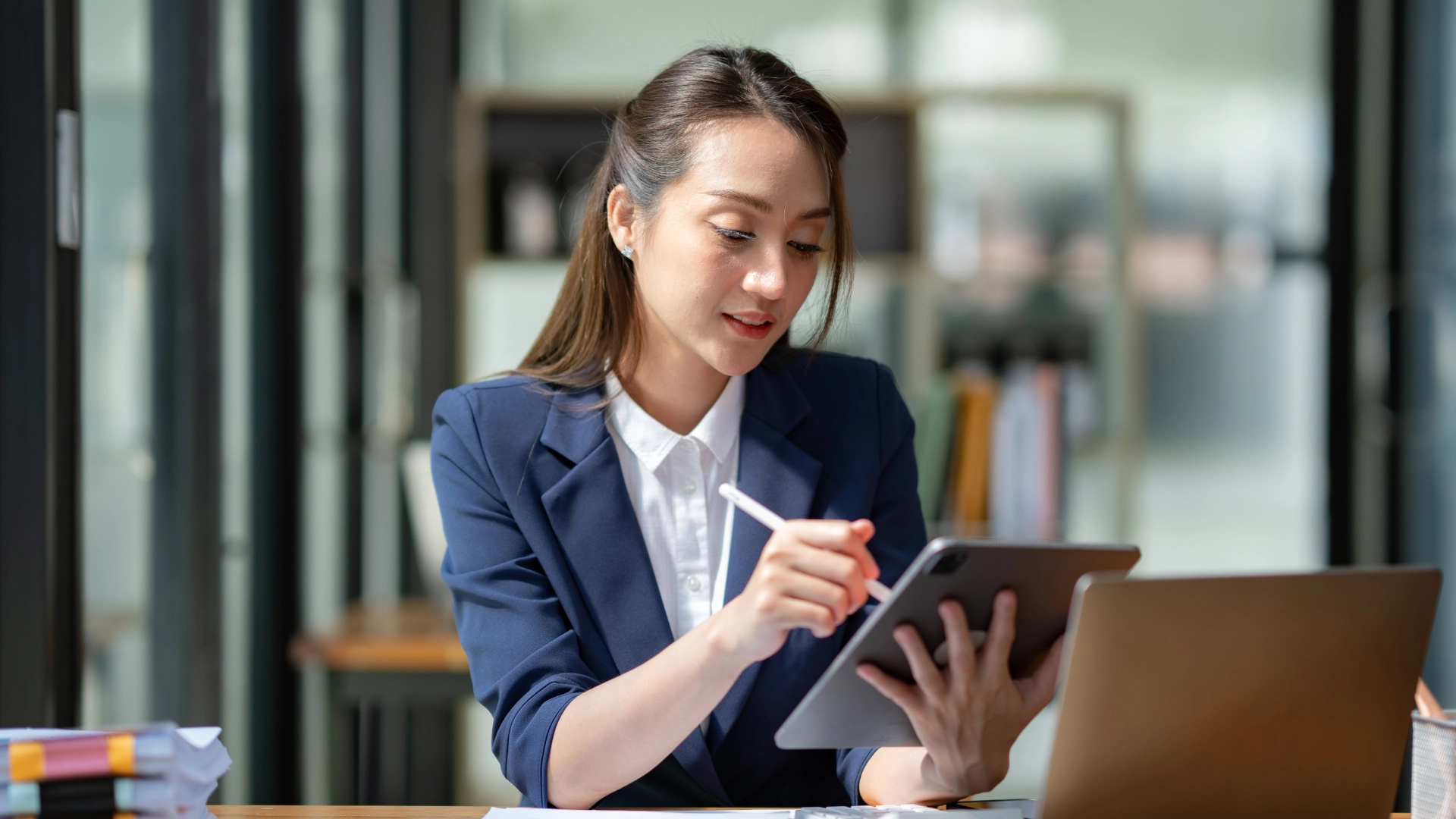 The woman, dressed in a blue blazer, is using a stylus to work on a tablet.