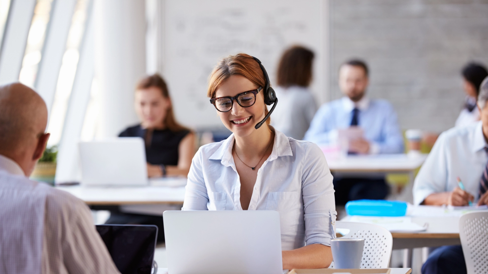 Smiling customer service representative with glasses wearing a headset and working on a laptop in a modern office.