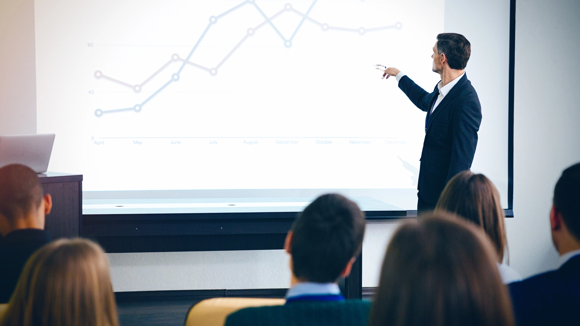 Man in a suit giving a presentation to an audience, pointing to a line graph on a screen.