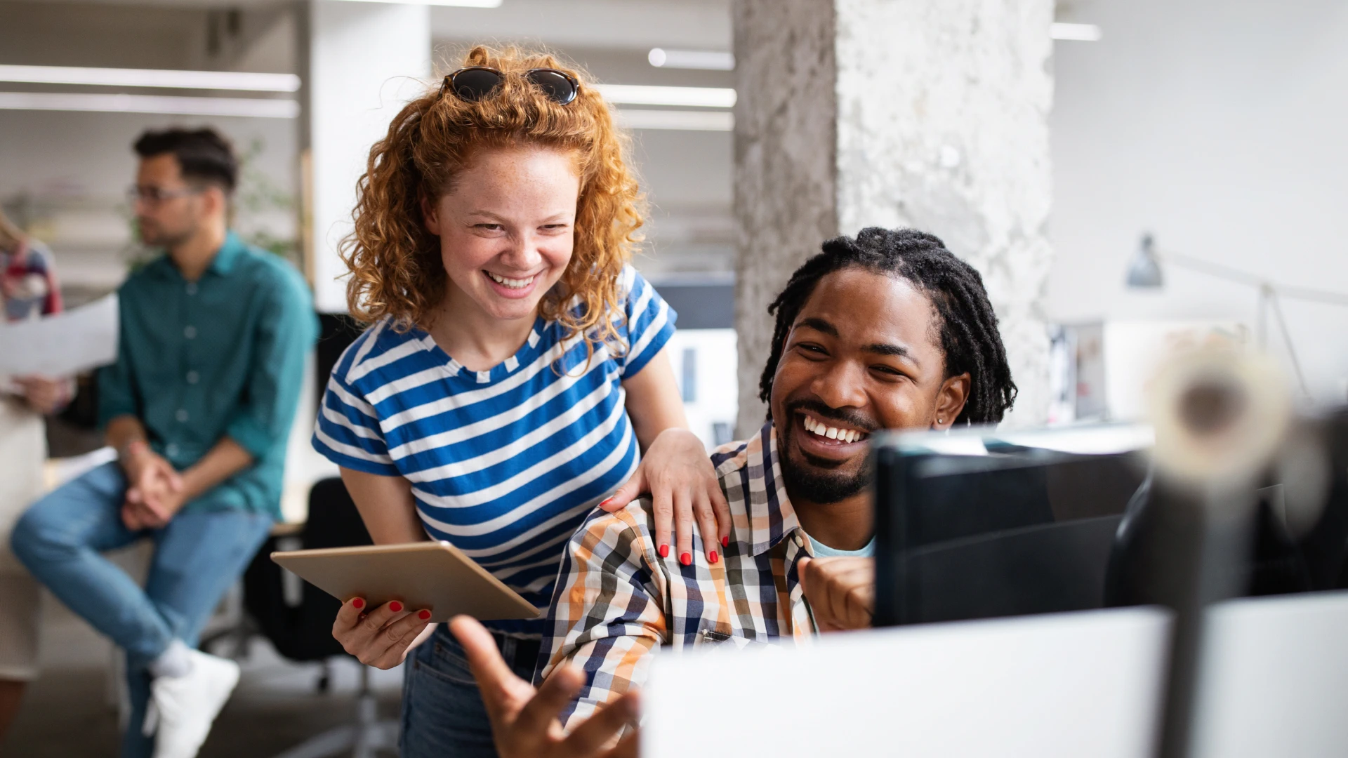 Cheerful colleagues collaborating at a computer, with woman holding a tablet and laughing with her coworker in a bright open office.