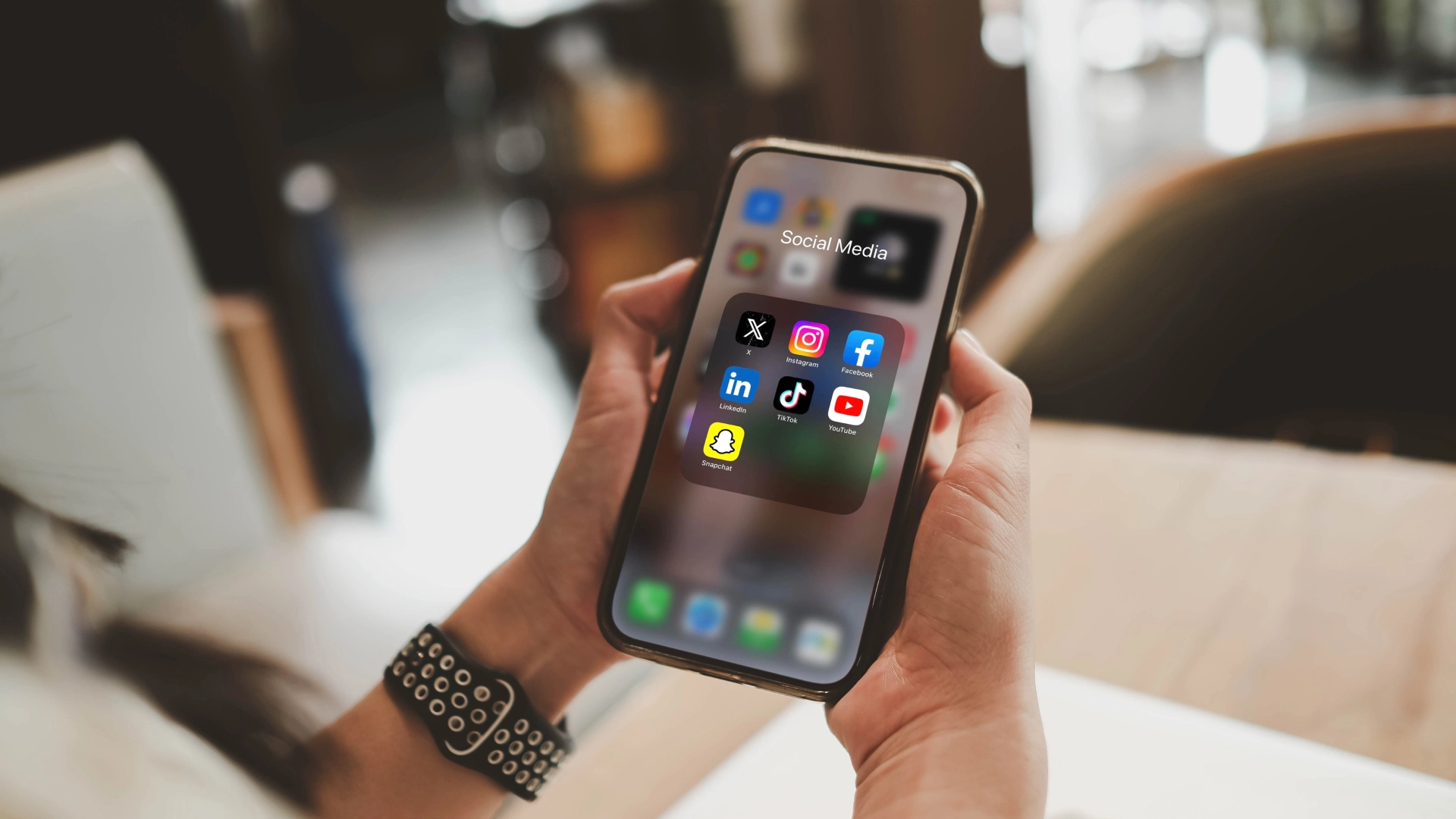 Person holding a smartphone displaying a folder of social media apps, including X, Instagram, Facebook, LinkedIn, TikTok, and Snapchat.